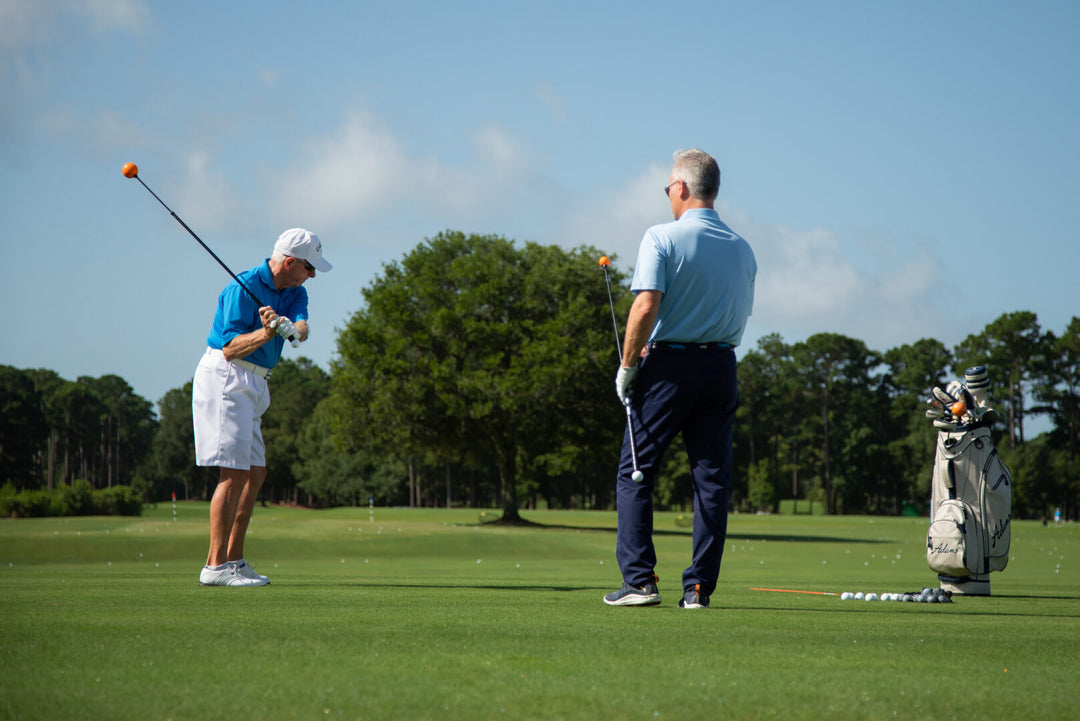 Two older men on a sunny golf course; one swings a club as the other watches, with a golf bag to the right.