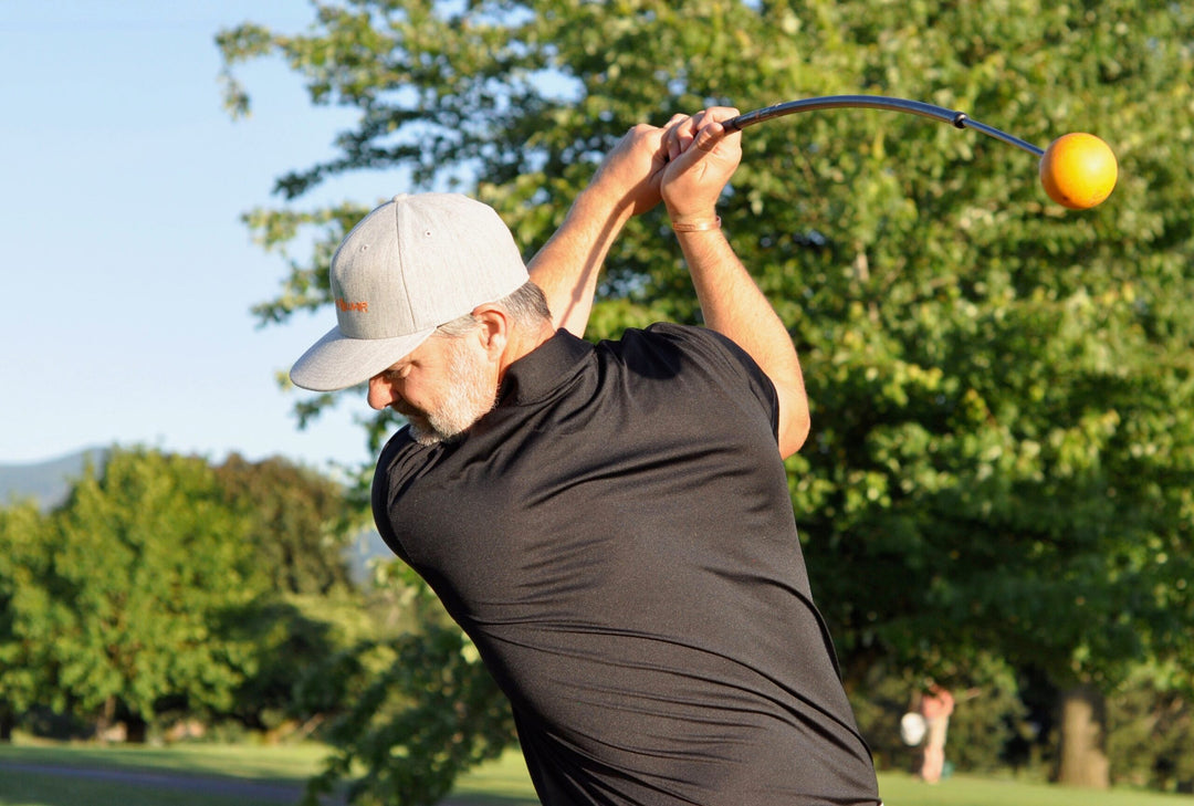 Man wearing a gray cap and black shirt swings a curved rod with an orange ball at the end in a park setting.