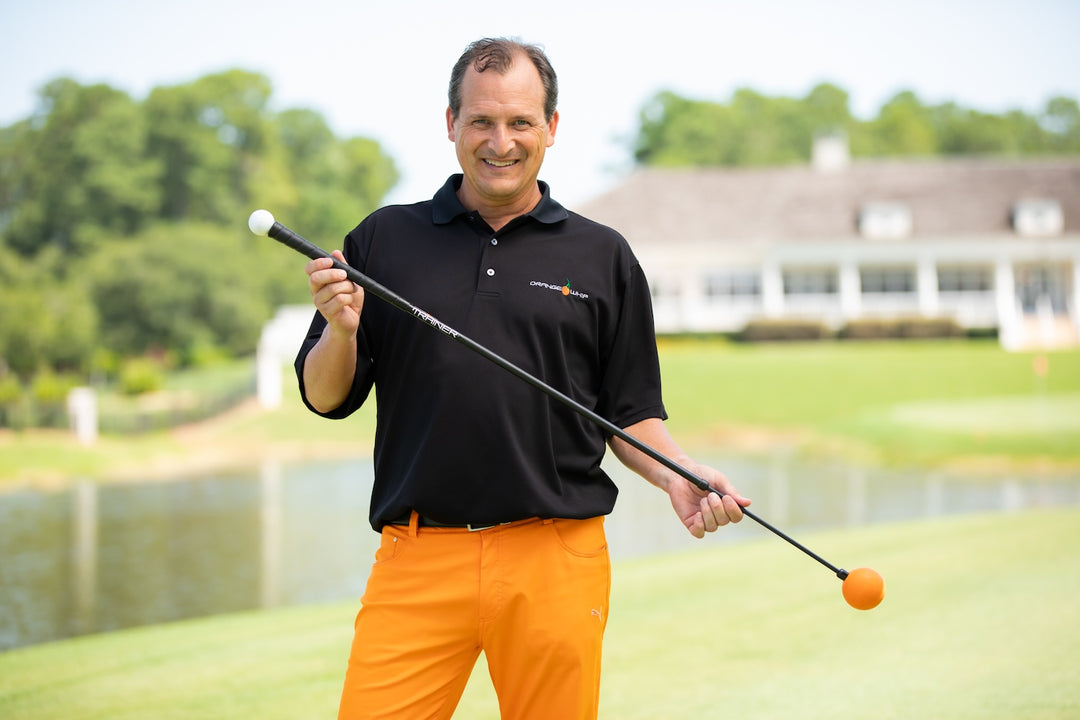 Smiling man in a black polo and orange pants holds a golf training stick on a sunny course with a water hazard in the background.