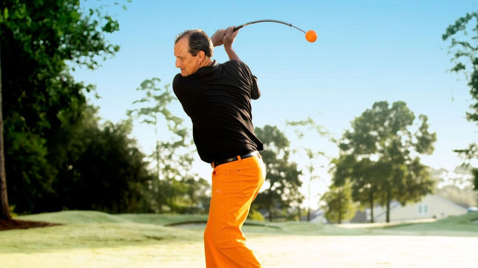 A man in a black polo shirt and bright orange pants swings a golf club on a sunny golf course, with trees and blue sky in the background.