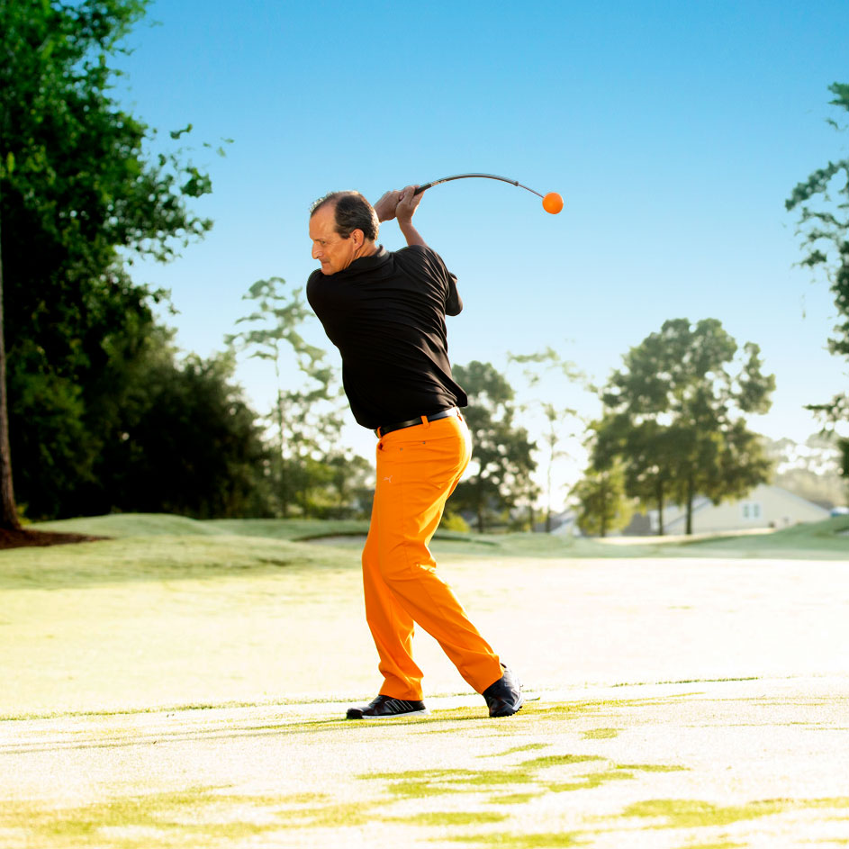 A man on a golf course wearing a black polo shirt and bright orange pants, captured mid-swing with a golf ball visible in the air.