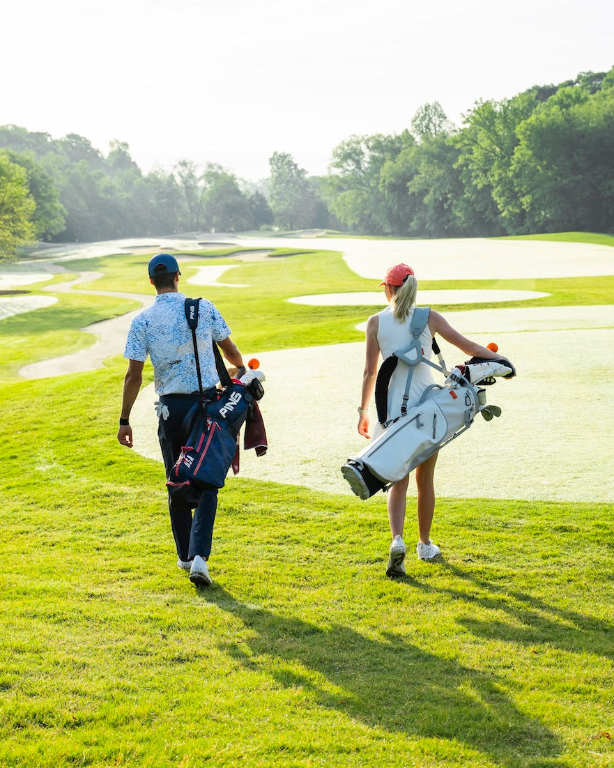 A man and a woman walk across a golf course carrying golf bags. The man wears a light blue patterned short-sleeve shirt, dark pants, and a blue cap, while the woman wears a sleeveless white top, a red cap, and carries a white golf bag.