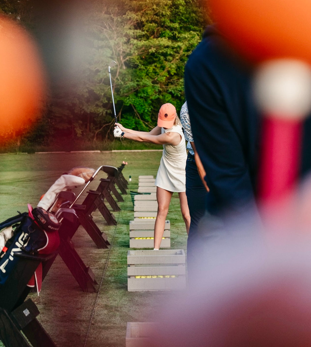 A woman in a white dress swings a golf club at a driving range, with other players and golf bags visible in the background and foreground.