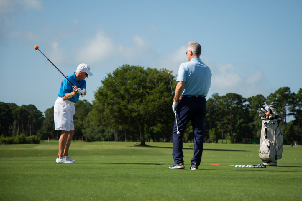 Two older men on a sunny golf course; one swings a club as the other watches, with a golf bag to the right.